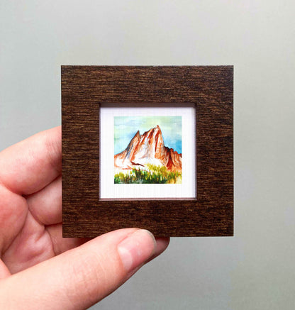 A hand is holding a small, square-shaped wooden frame containing a watercolor painting of a mountain landscape.