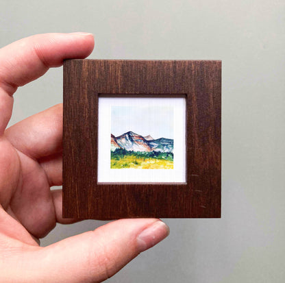 A hand is holding a small wooden frame containing a watercolor painting of a mountain landscape.