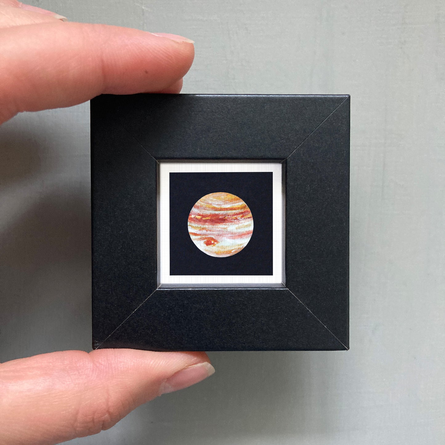 A person's hand is holding a small black square frame containing a miniature representation of the planet Jupiter.