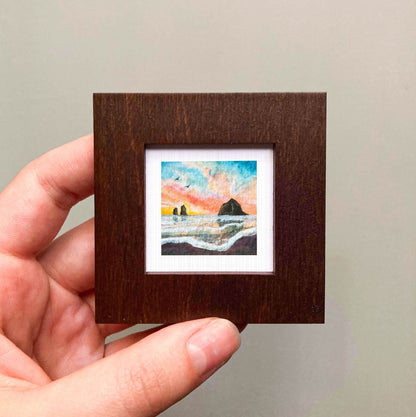 A hand is holding a small, square-shaped wooden frame containing a painting of a seascape with a rock formation and a sunset in the background.