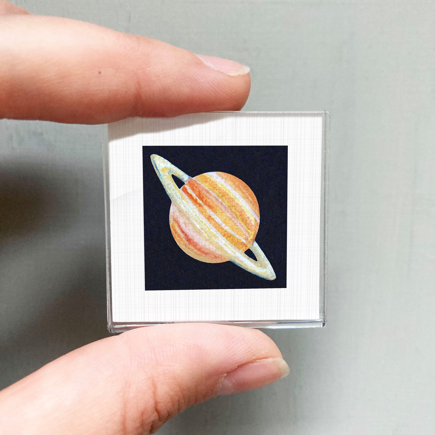 A person's hand is holding a small square picture of a planet with rings.