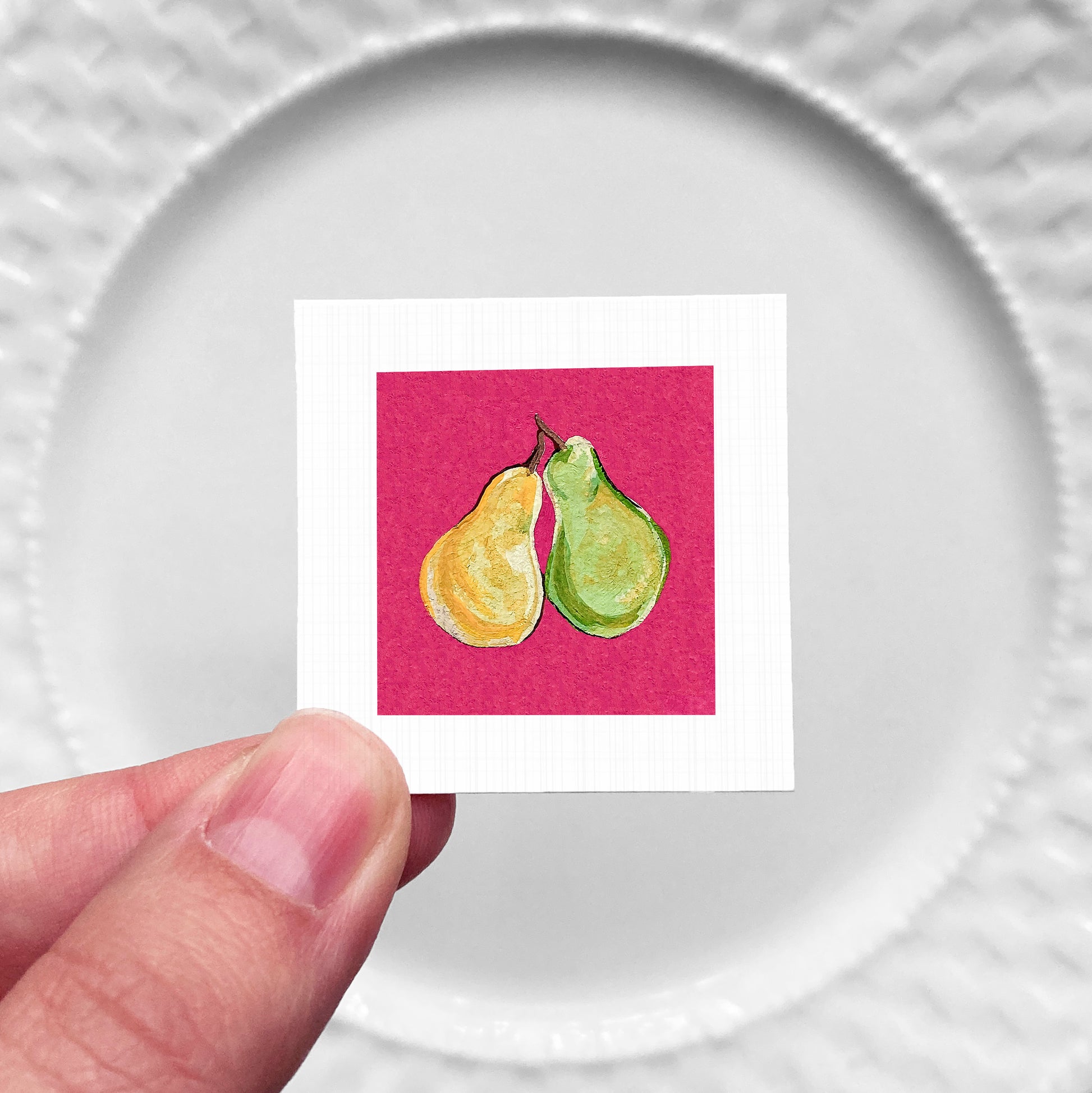A hand is holding a small square with a painting of two pears on a pink background.