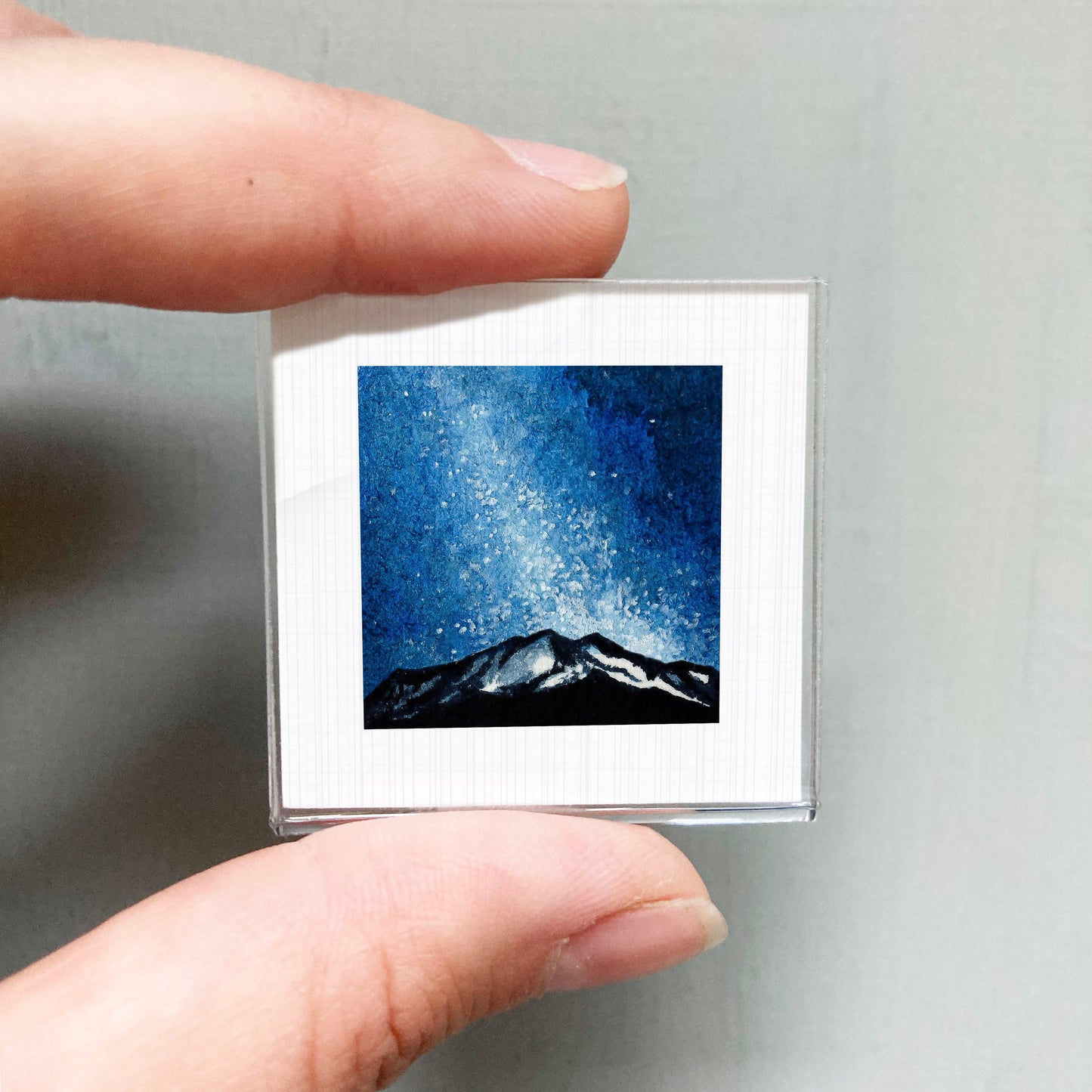 A person's hand holding a small square picture of a mountain landscape under a starry night sky.