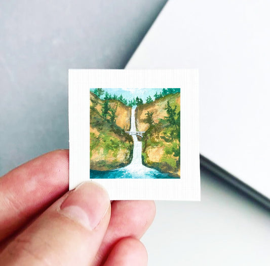 A hand holding a small square picture depicting a waterfall surrounded by lush greenery.