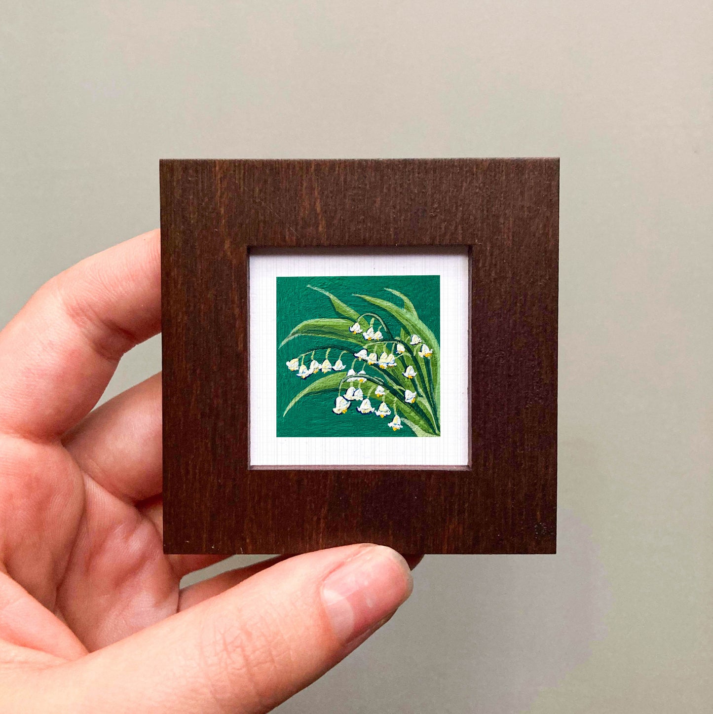 A hand is holding a small wooden frame containing a framed painting of lily of the valley flowers.