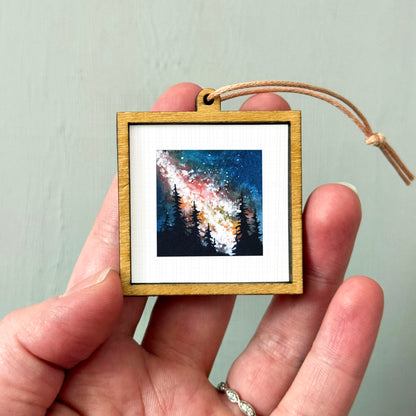 A hand holding a small wooden frame containing a painting of a night sky with stars and trees.