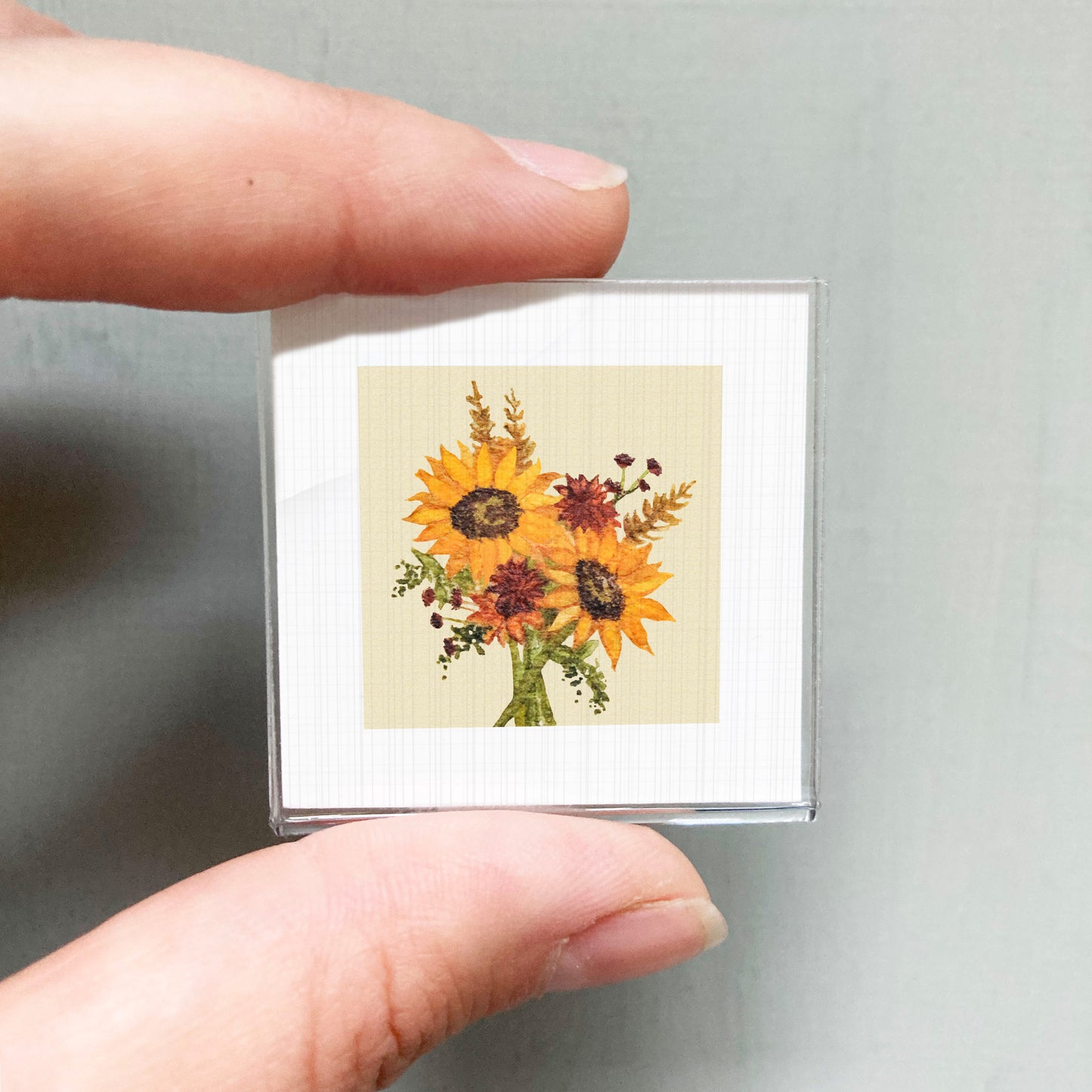 A person's hand holding a small square picture of a bouquet of yellow sunflowers.