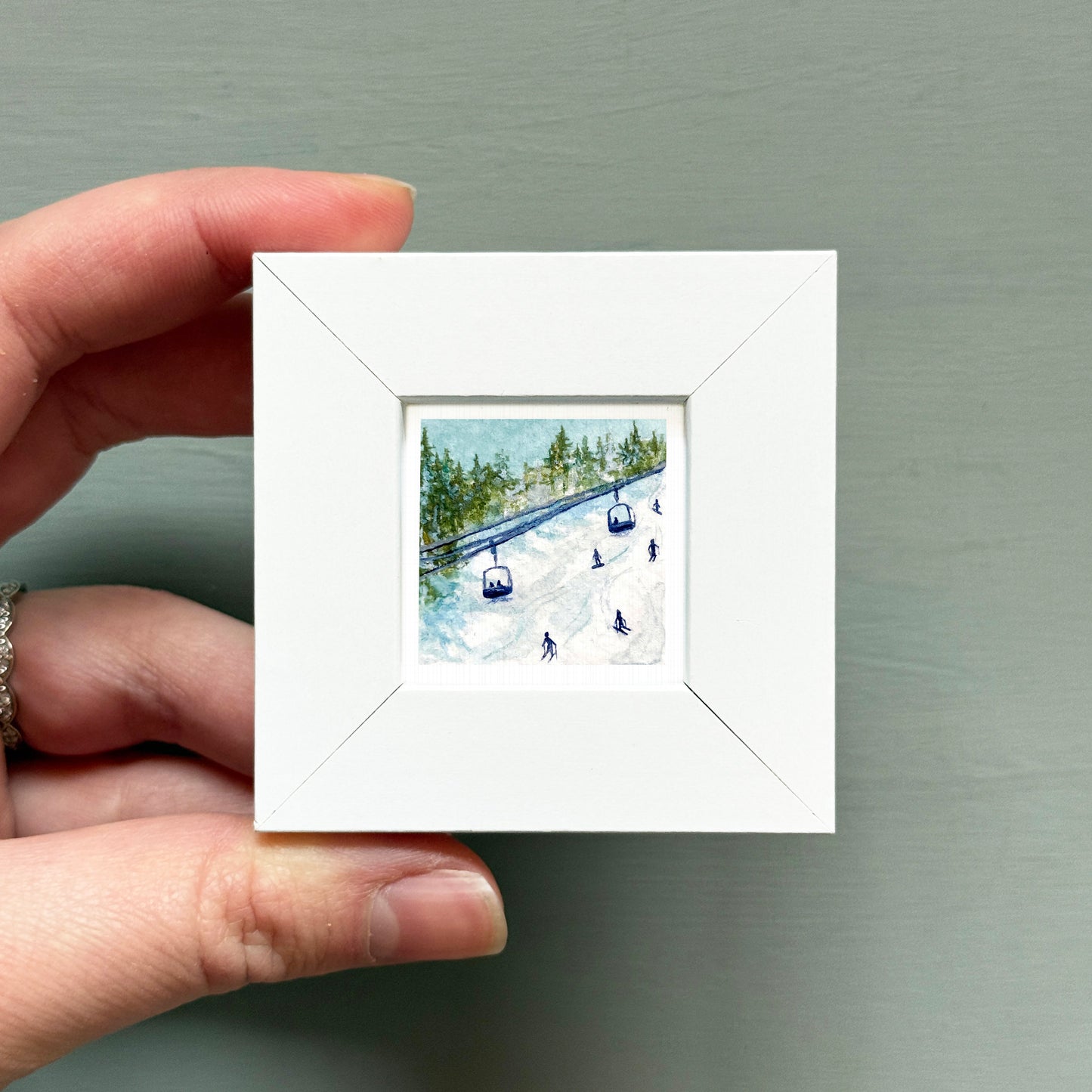 A hand is holding a small white square frame containing a painting of a ski lift and people skiing.