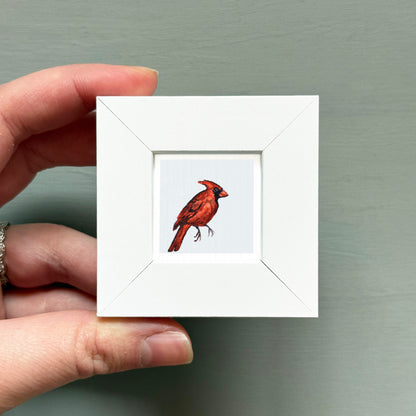 A hand is holding a small white frame containing a framed image of a red cardinal bird.