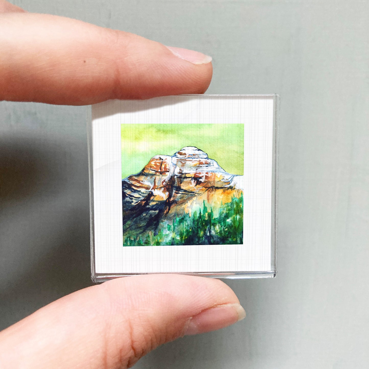 A person's hand holding a small, framed painting of a mountain landscape.