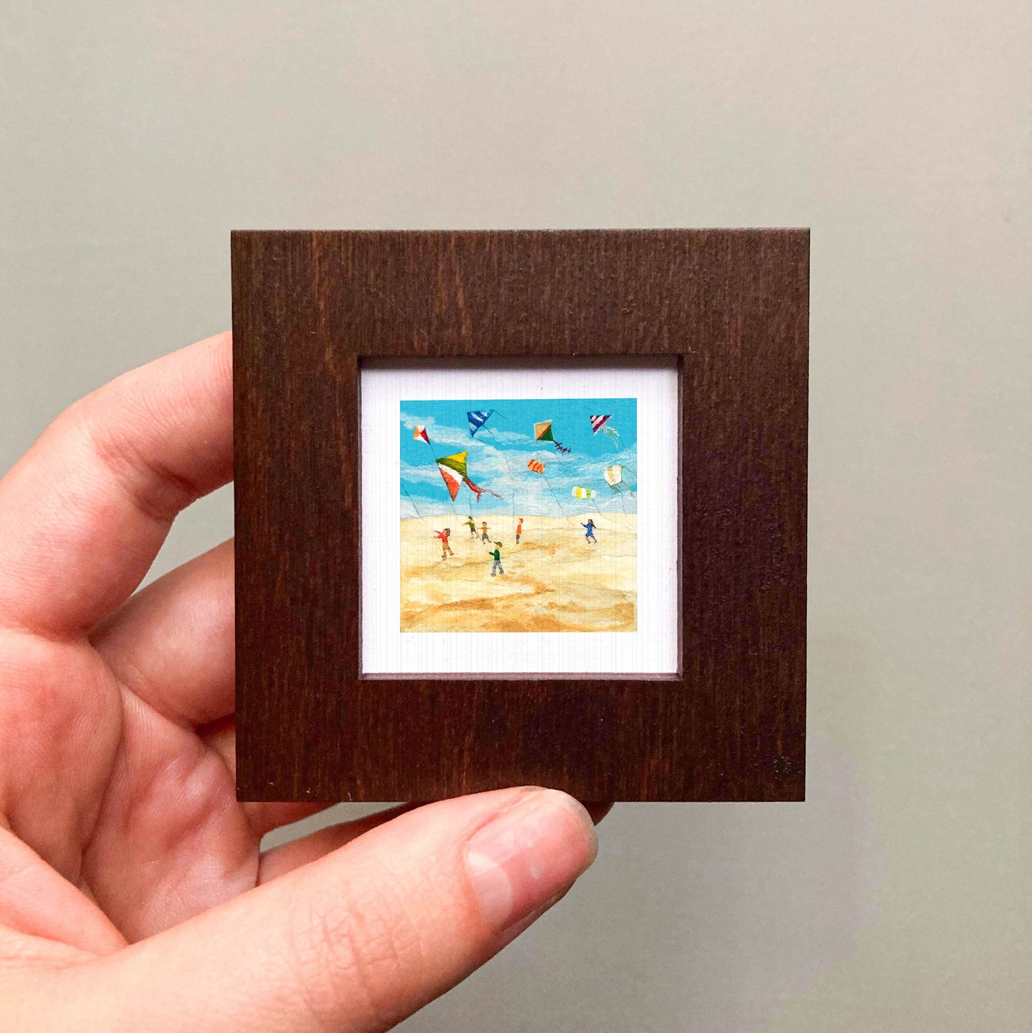 A hand is holding a small, square-shaped picture frame containing a painting of people flying kites on a beach.