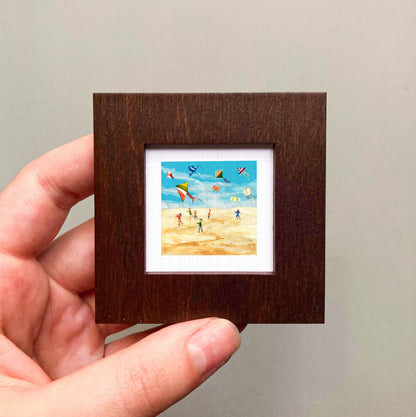 A hand is holding a small, square-shaped picture frame containing a painting of people flying kites on a beach.