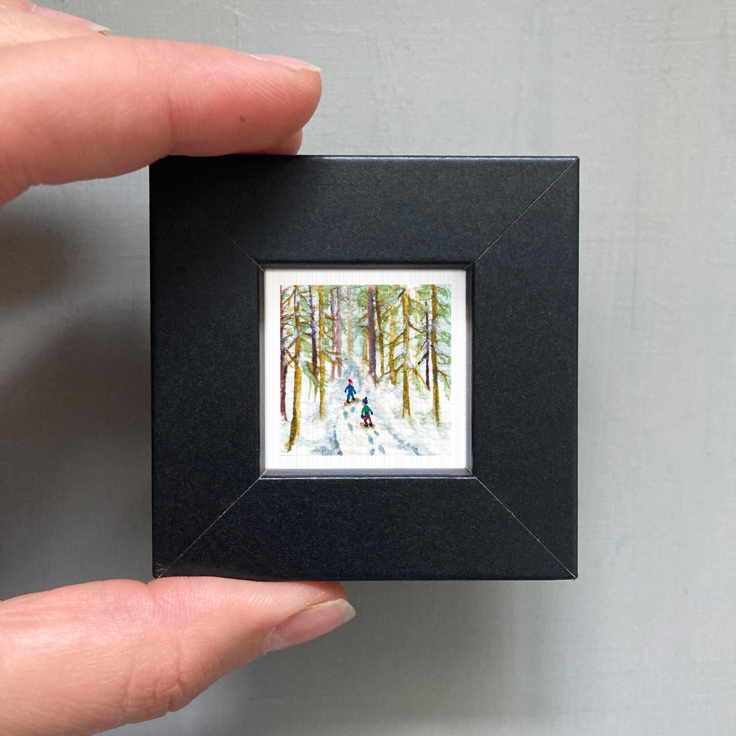 A person's hand is holding a small black frame containing a miniature painting of two people skiing in a snowy forest.