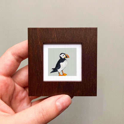 A person's hand is holding a small wooden frame containing a framed image of a puffin bird.