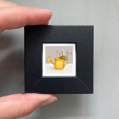 A hand is holding a small black frame containing a framed painting of a yellow watering can with flowers.