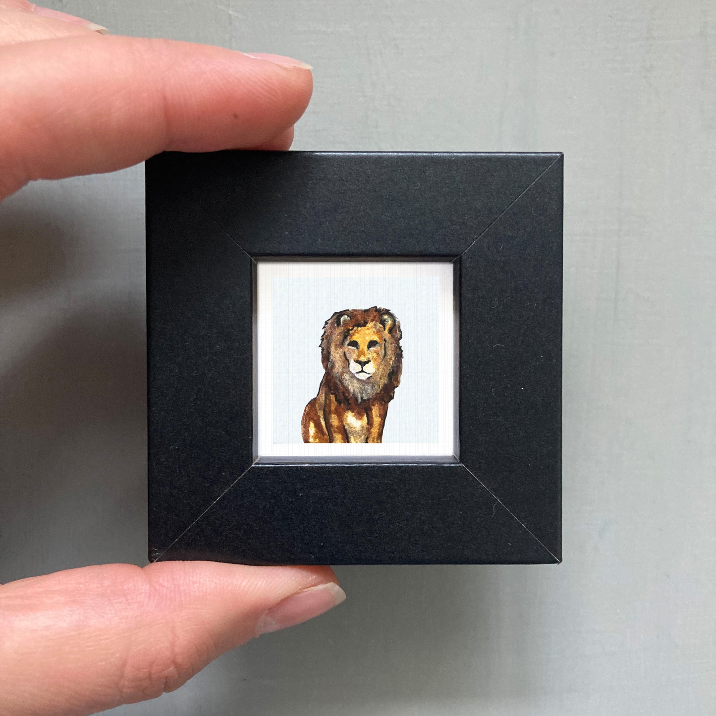 A person's hand is holding a small black picture frame containing a watercolor painting of a lion.