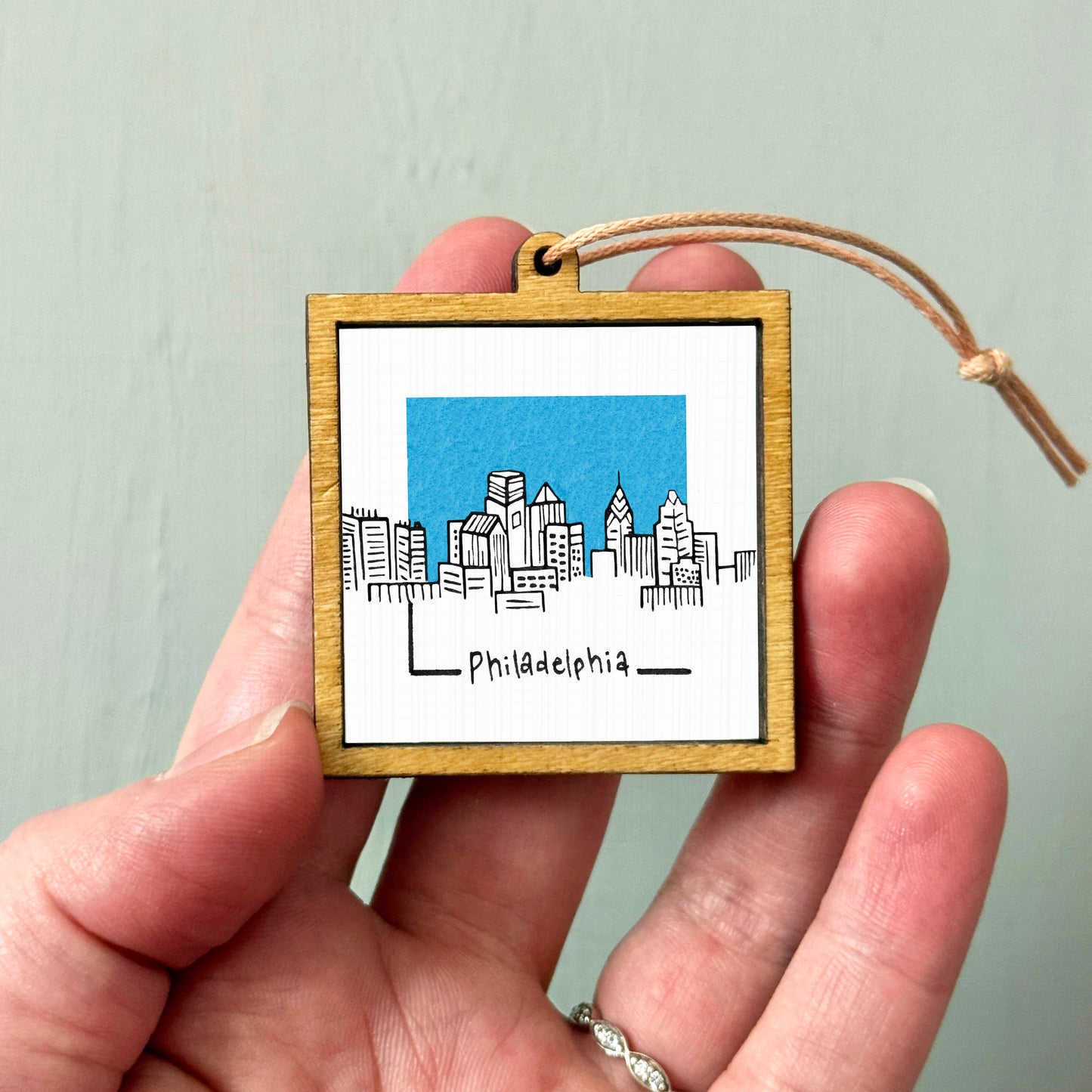 A hand holding a small wooden frame with a picture of a city skyline and the word "Philadephia" written on it.