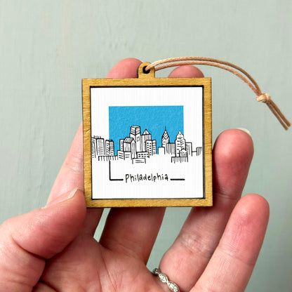 A hand holding a small wooden frame with a picture of a city skyline and the word "Philadephia" written on it.