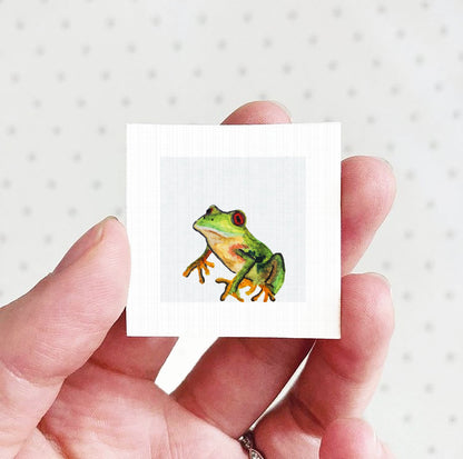 A person's hand holding a small square picture of a green frog against a white background.
