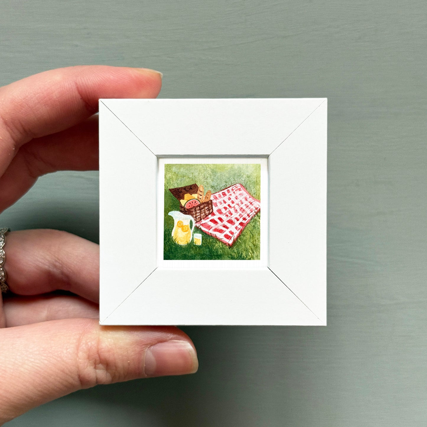 A hand is holding a small white picture frame containing a painting of a picnic scene with a basket, blanket, and food items.