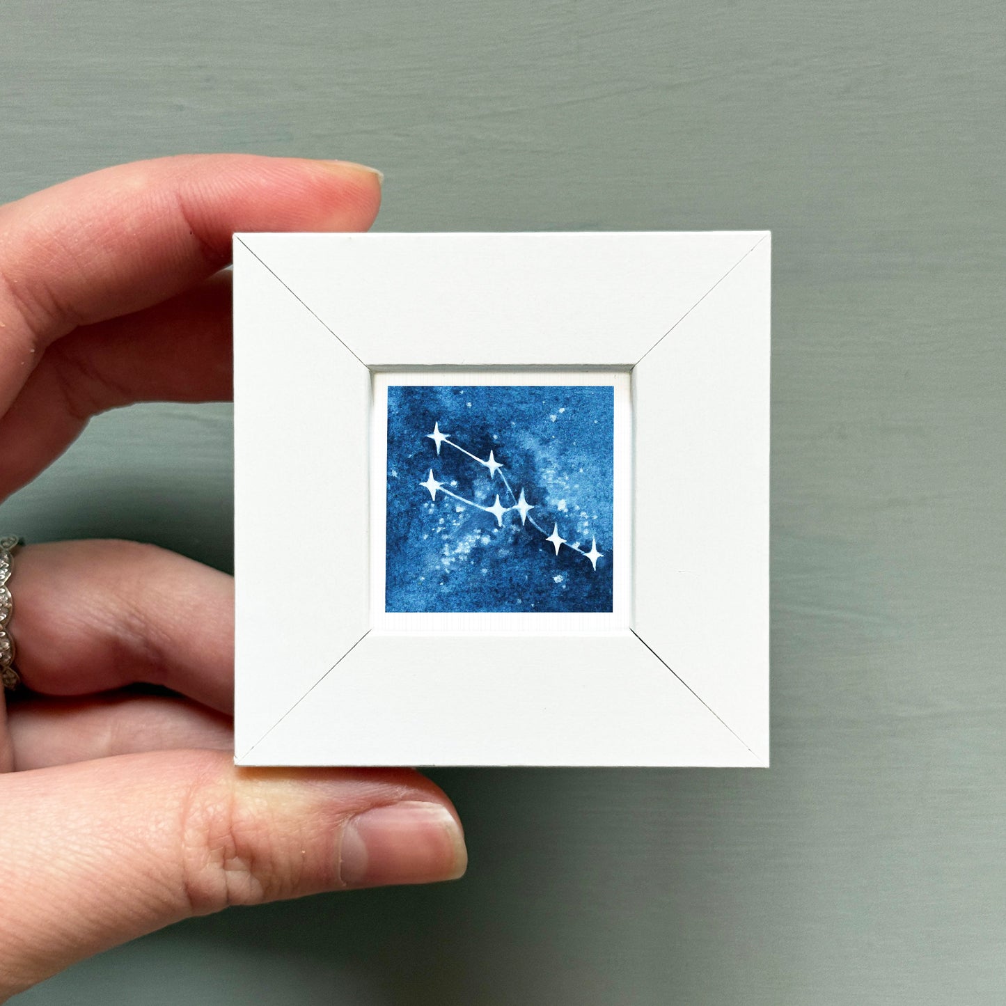 A hand is holding a small white square frame containing a blue and white starry night sky with a constellation.