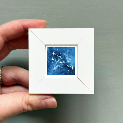 A hand is holding a small white square frame containing a blue and white starry night sky with a constellation.