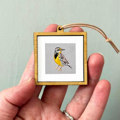 A hand holding a small wooden frame containing a framed image of a bird.
