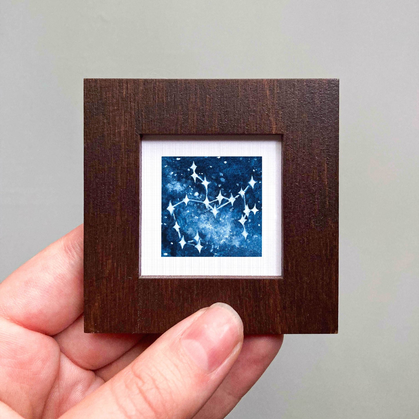 A hand is holding a small wooden frame containing a framed image of a starry night sky.