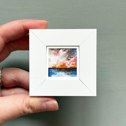 A hand is holding a small white square frame containing a painting of birds flying over a body of water.