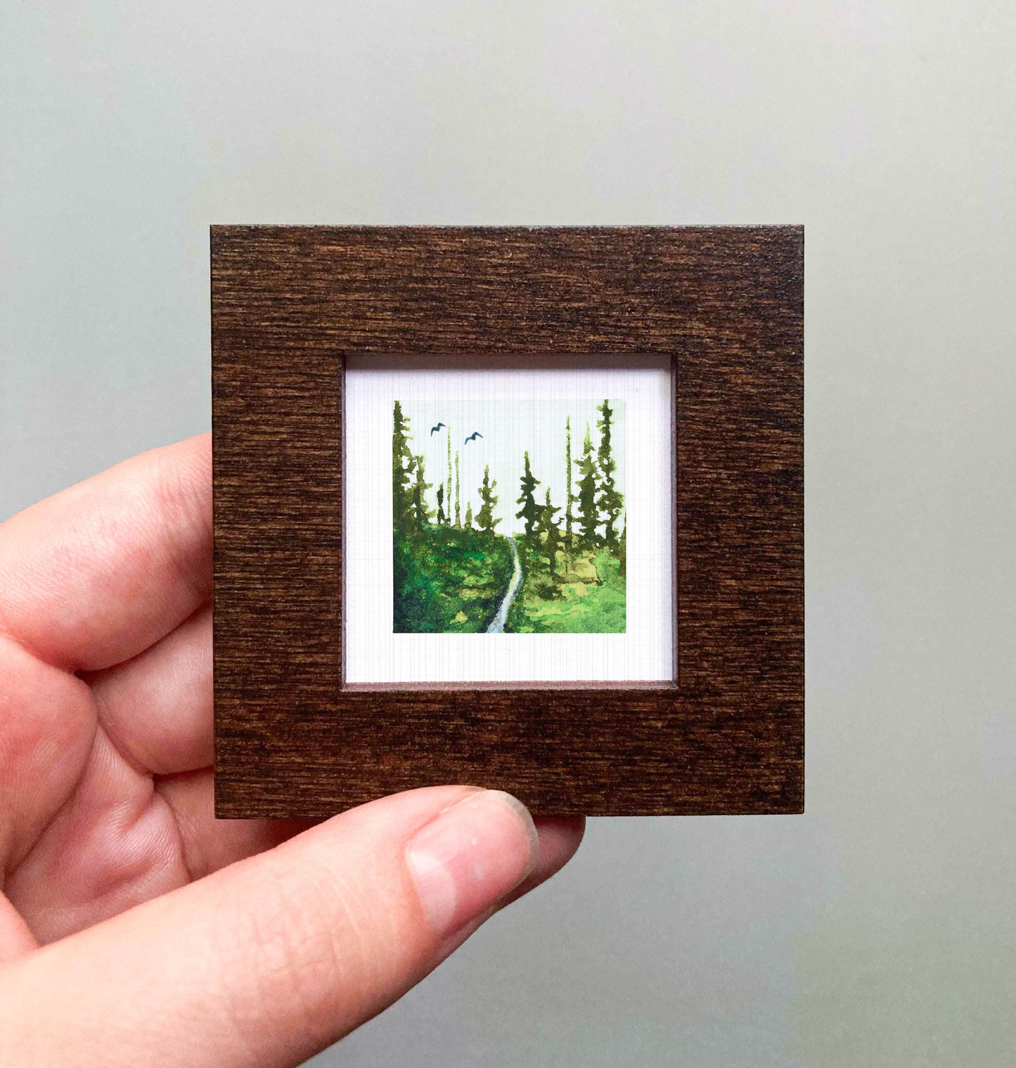A hand is holding a small, square-shaped picture frame containing a painting of a forest scene.
