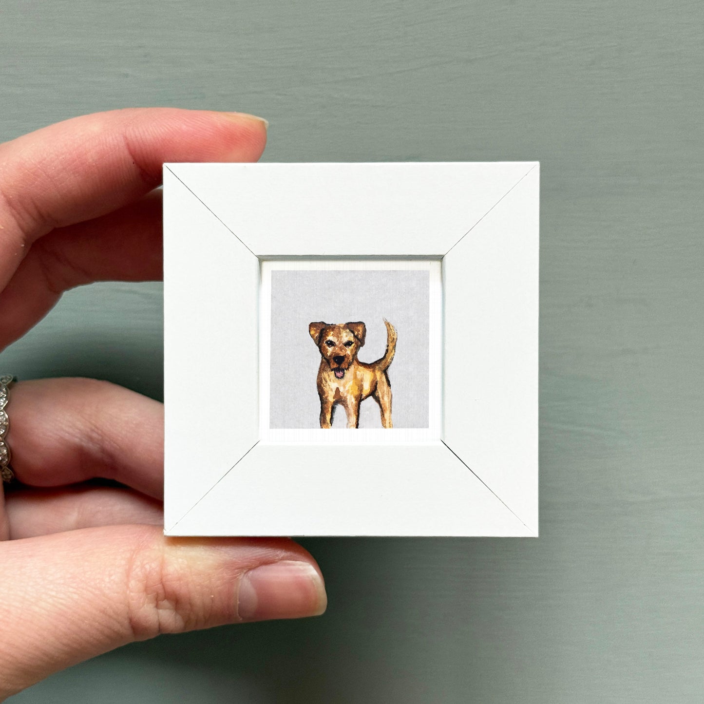 A hand is holding a small white picture frame containing a painting of a dog.