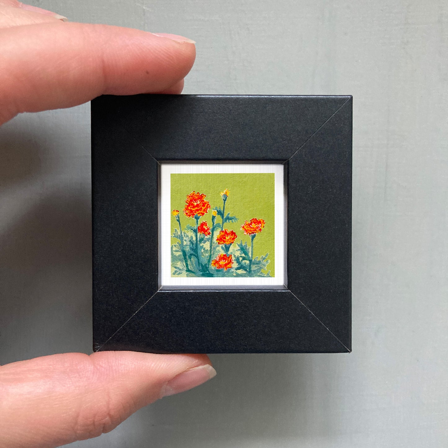 A person's hand holding a small black frame containing a framed painting of red flowers against a green background.