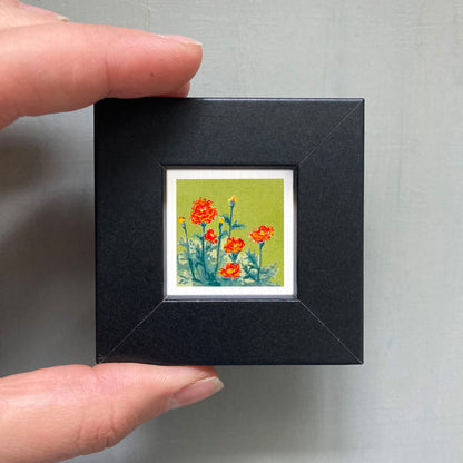 A person's hand holding a small black frame containing a framed painting of red flowers against a green background.
