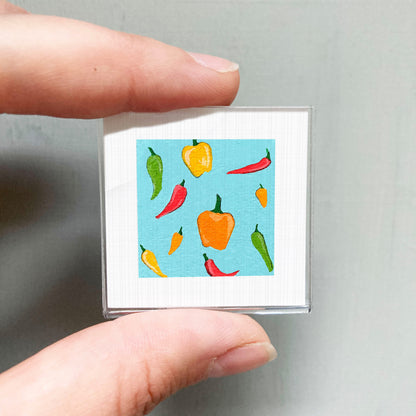 A person's hand is holding a small square with a vibrant illustration of various colorful peppers on a blue background.