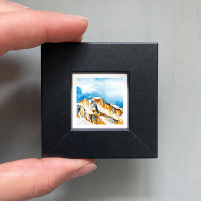 A person's hand holding a small black frame containing a watercolor painting of a mountain landscape.
