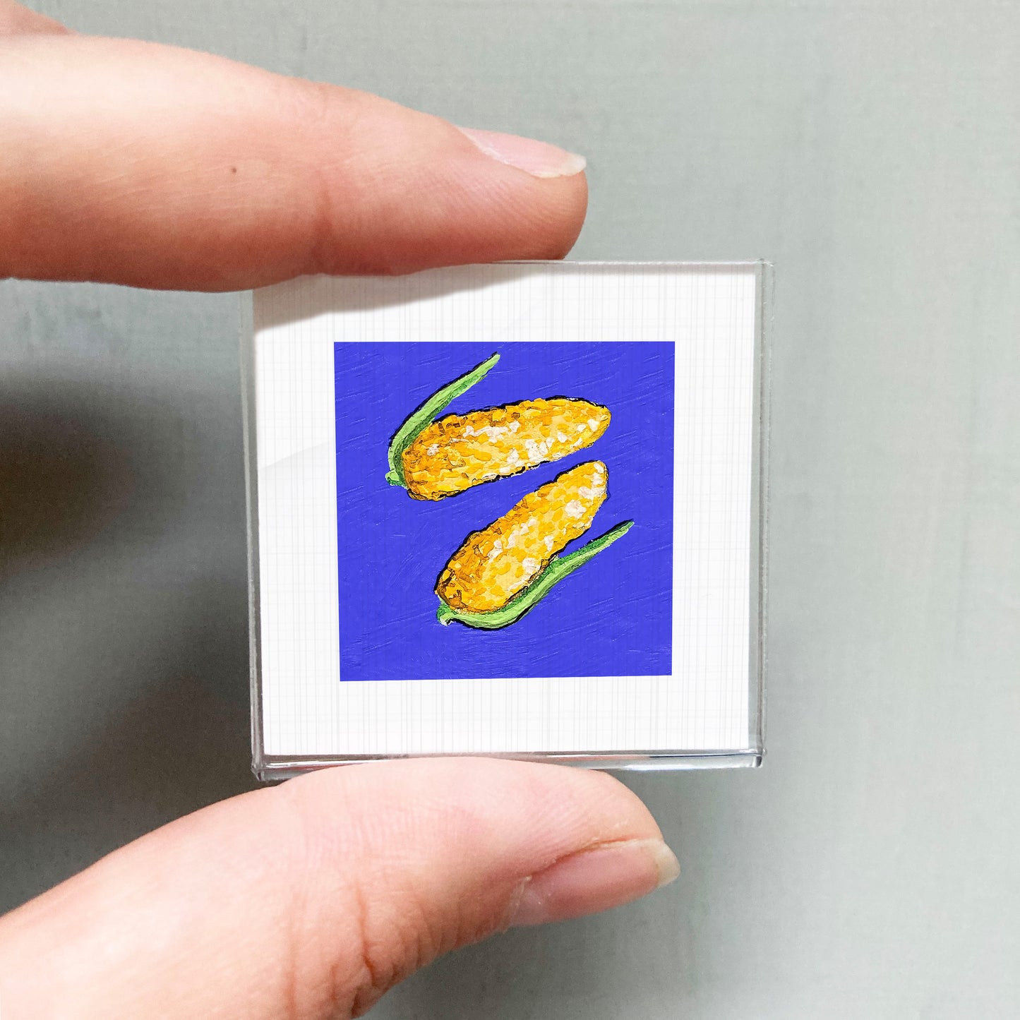 A person's hand is holding a small square with a vibrant painting of two yellow corn cobs against a blue background.