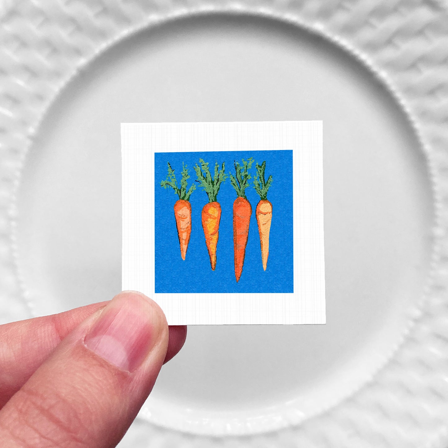 A hand is holding a small square with a vibrant illustration of four carrots against a blue background.