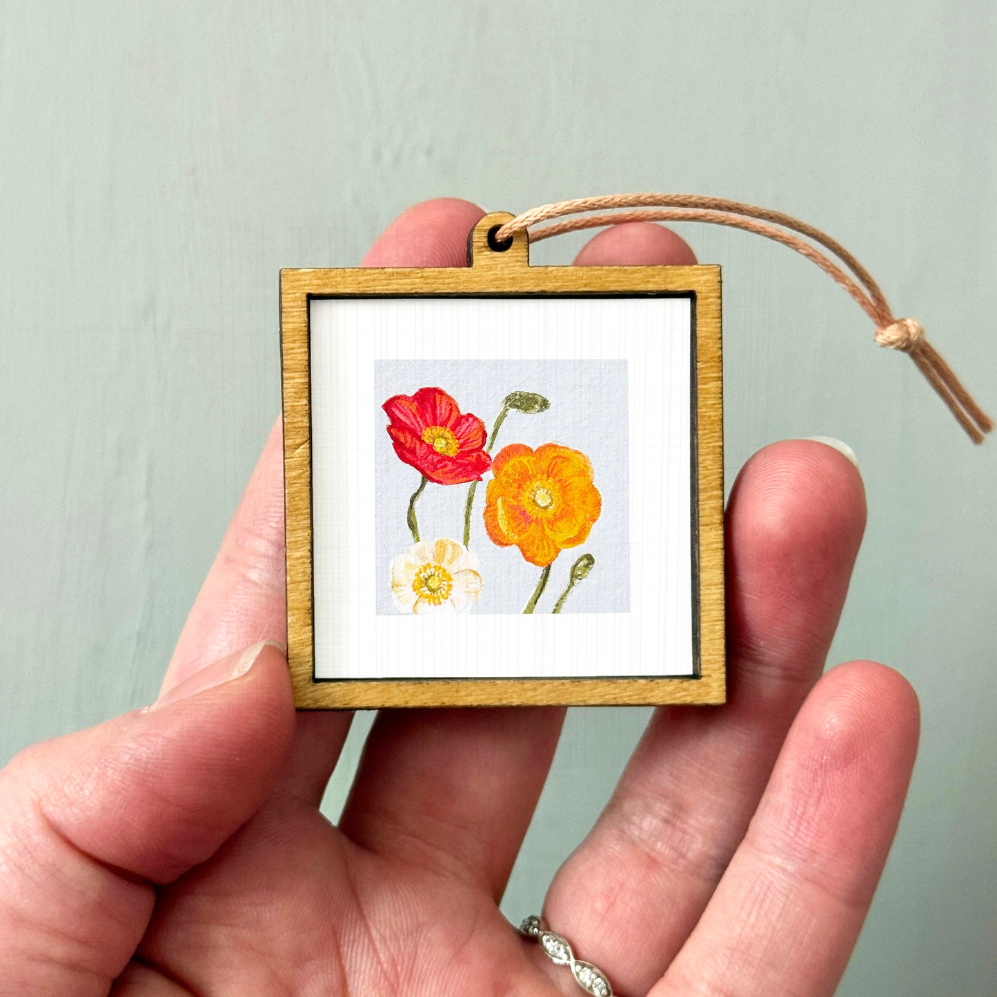 A hand holding a small wooden frame containing a picture of three flowers - two red and one yellow.