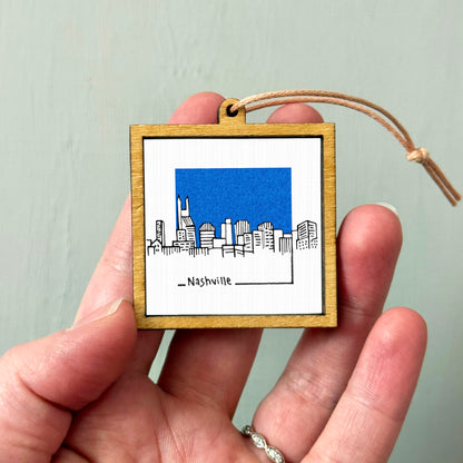 A hand holding a small wooden frame with a picture of a city skyline, specifically Nashville, inside it.