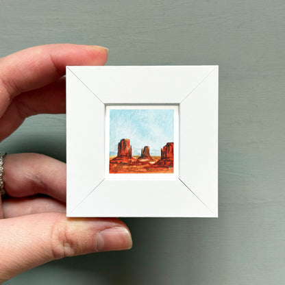 A hand is holding a small white square frame containing a miniature painting of a desert landscape with three rock formations.