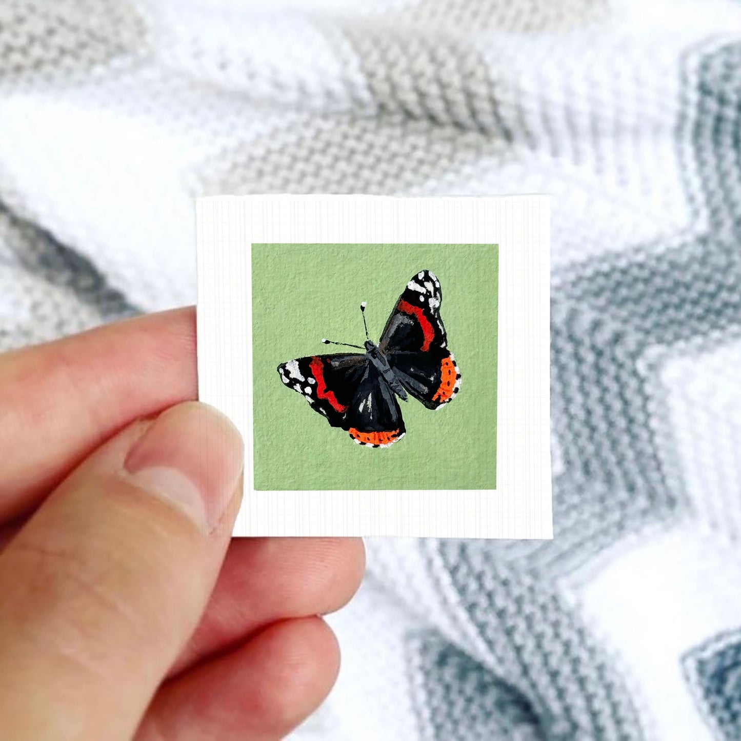 A hand is holding a small square card with a picture of a colorful butterfly on it.