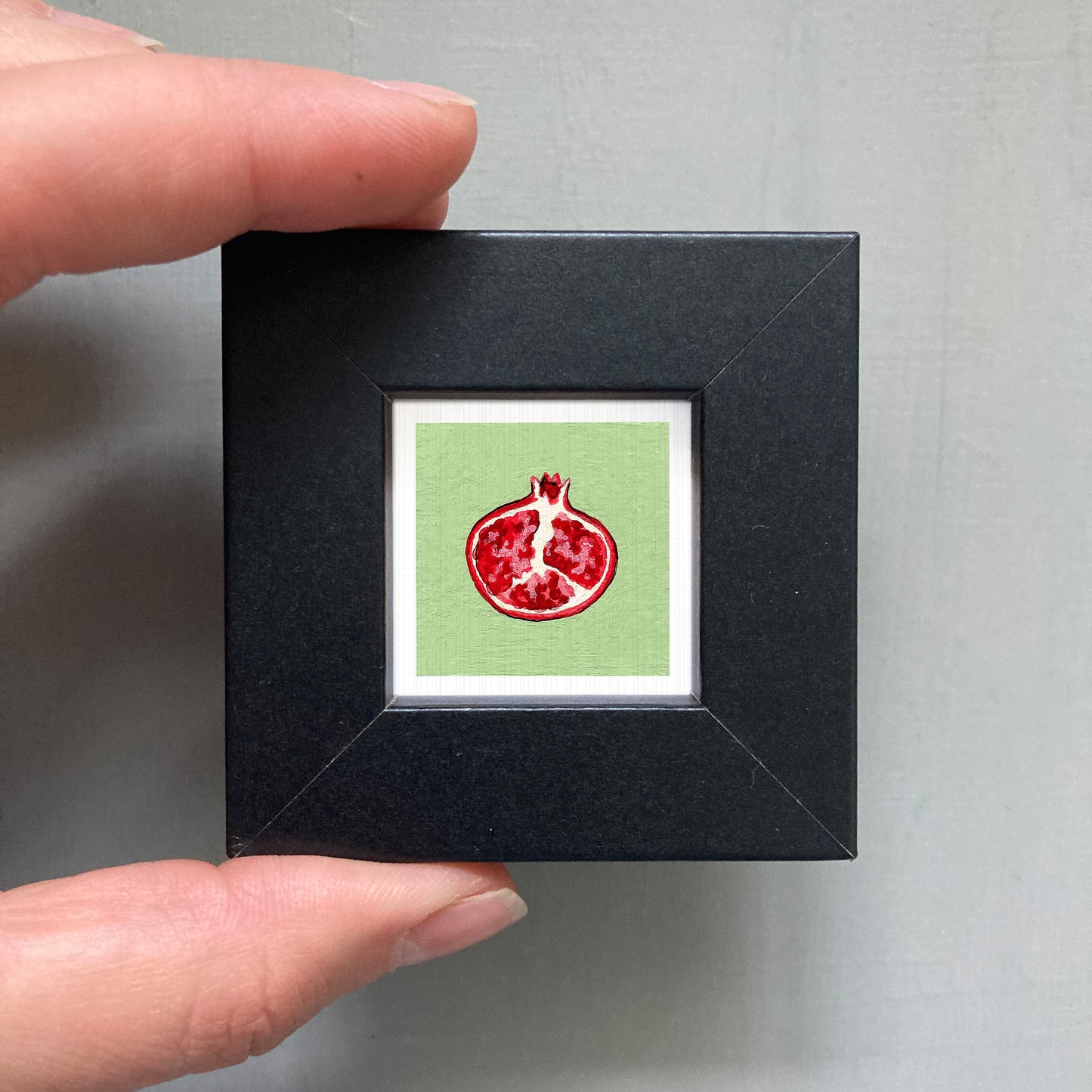 A person's hand holding a small black square frame containing a framed illustration of a pomegranate fruit.