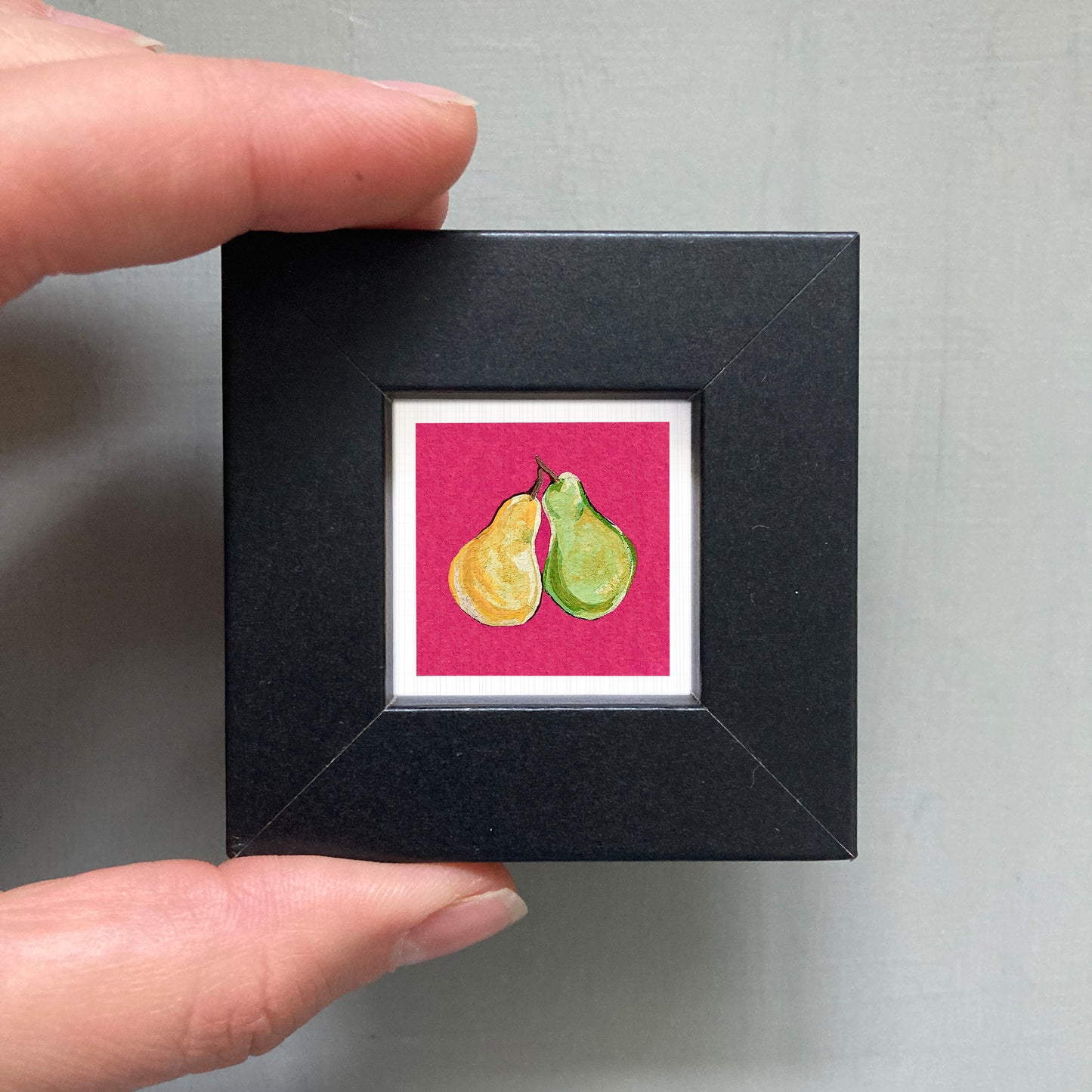 A hand is holding a small square picture frame containing a painting of two pears.