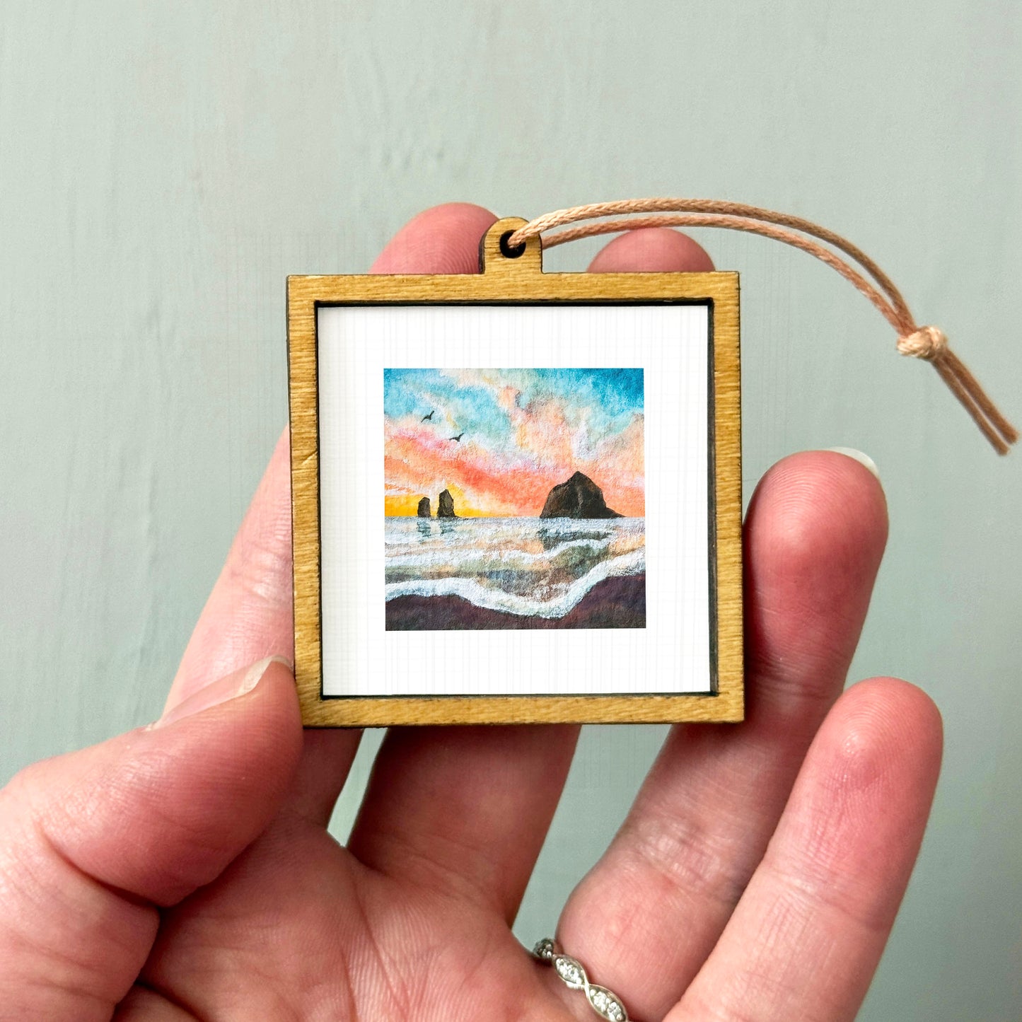 A hand holding a small wooden frame containing a painting of a beach scene with a sunset.