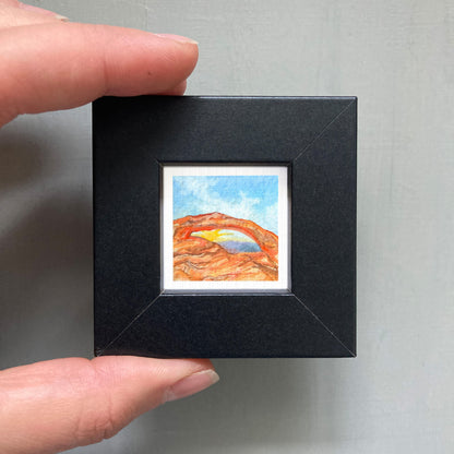 A person's hand holding a small black frame containing a watercolor painting of a desert landscape with a rainbow in the sky.