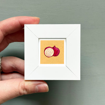 A hand is holding a small white square frame containing an illustration of a red onion.