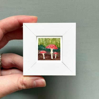 A hand is holding a small white frame containing a painting of three red mushrooms against a forest background.