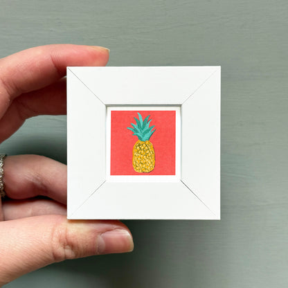 A hand is holding a small white picture frame containing a vibrant illustration of a pineapple.