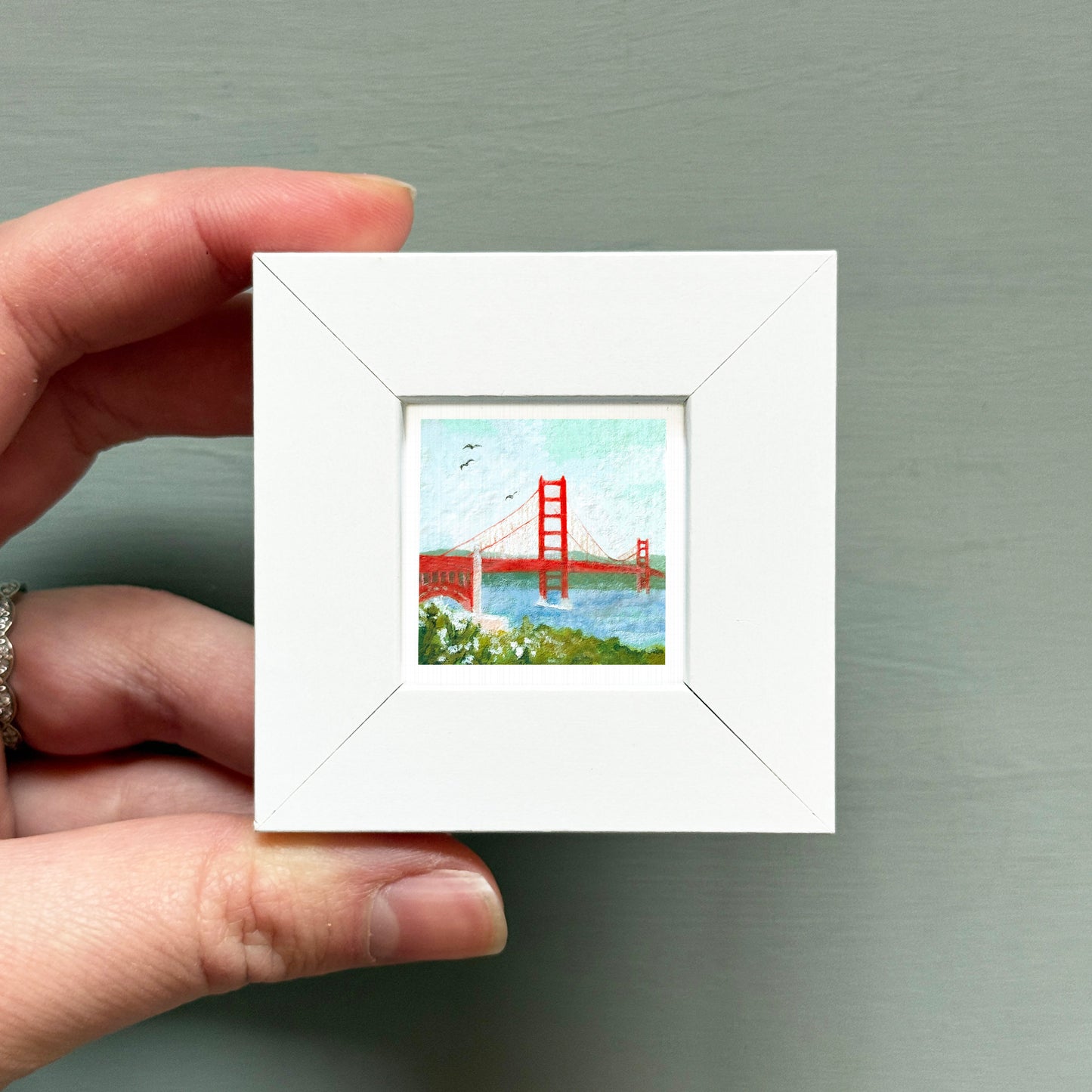 A hand is holding a small white square frame containing a painting of the Golden Gate Bridge.