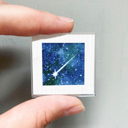 A hand is holding a small square picture of a starry night sky.